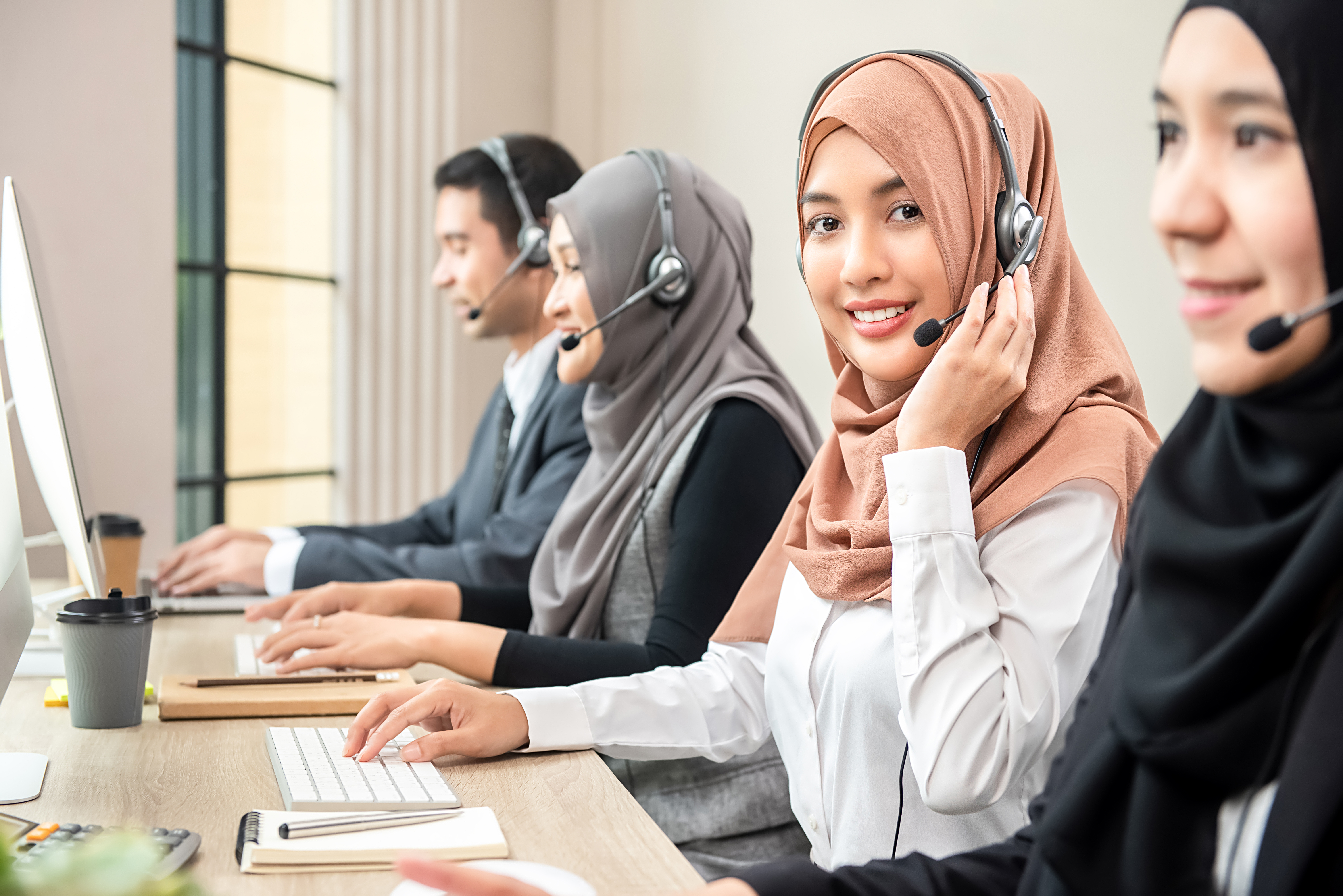 Friendly beautiful Asian muslim woman wearing microphone headset working as customer support operator with team in call center office